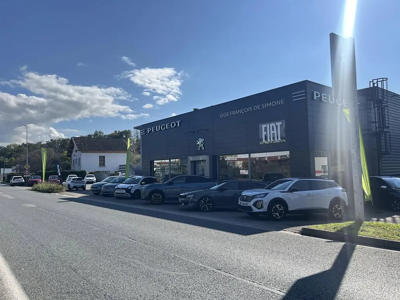 Façade d'une concession Peugeot et Fiat sous un ciel bleu, avec plusieurs voitures garées devant le bâtiment gris foncé.