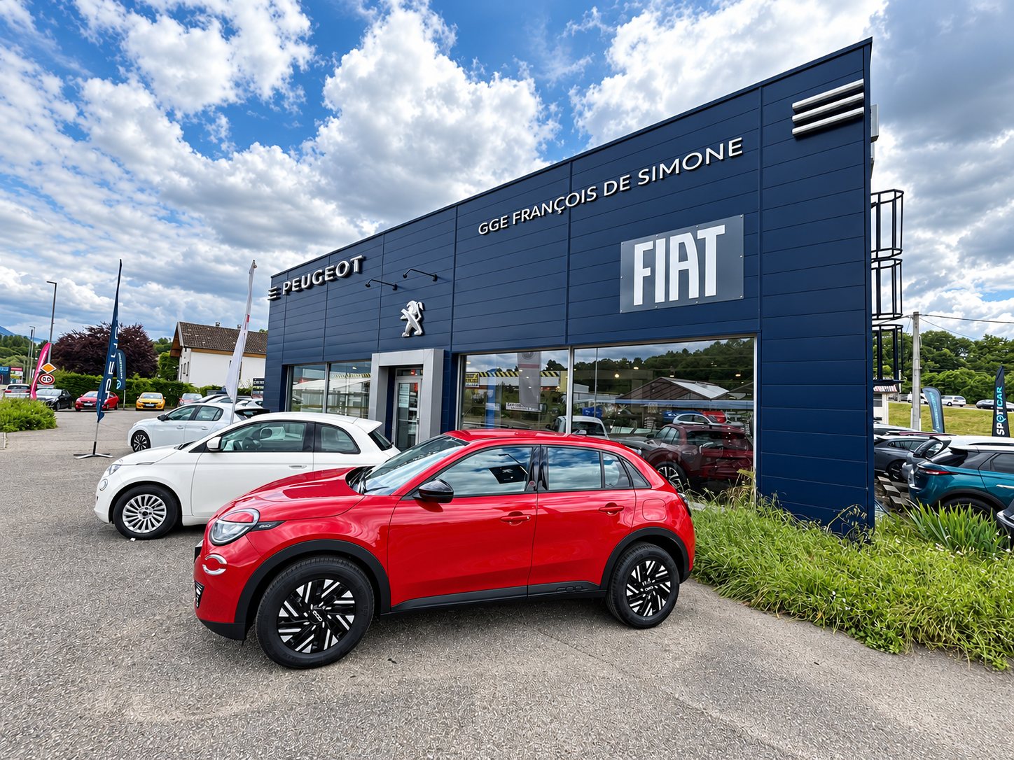 Façade bleu foncé d'une concession Peugeot et Fiat, avec une voiture rouge et une voiture blanche garées sur le parking devant le bâtiment.