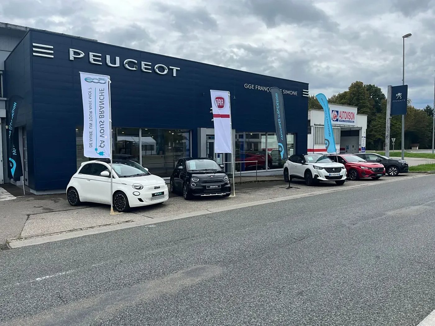 Façade bleue d'une concession Peugeot avec des Fiat 500 et des Peugeot garées devant, entourées de drapeaux publicitaires.
