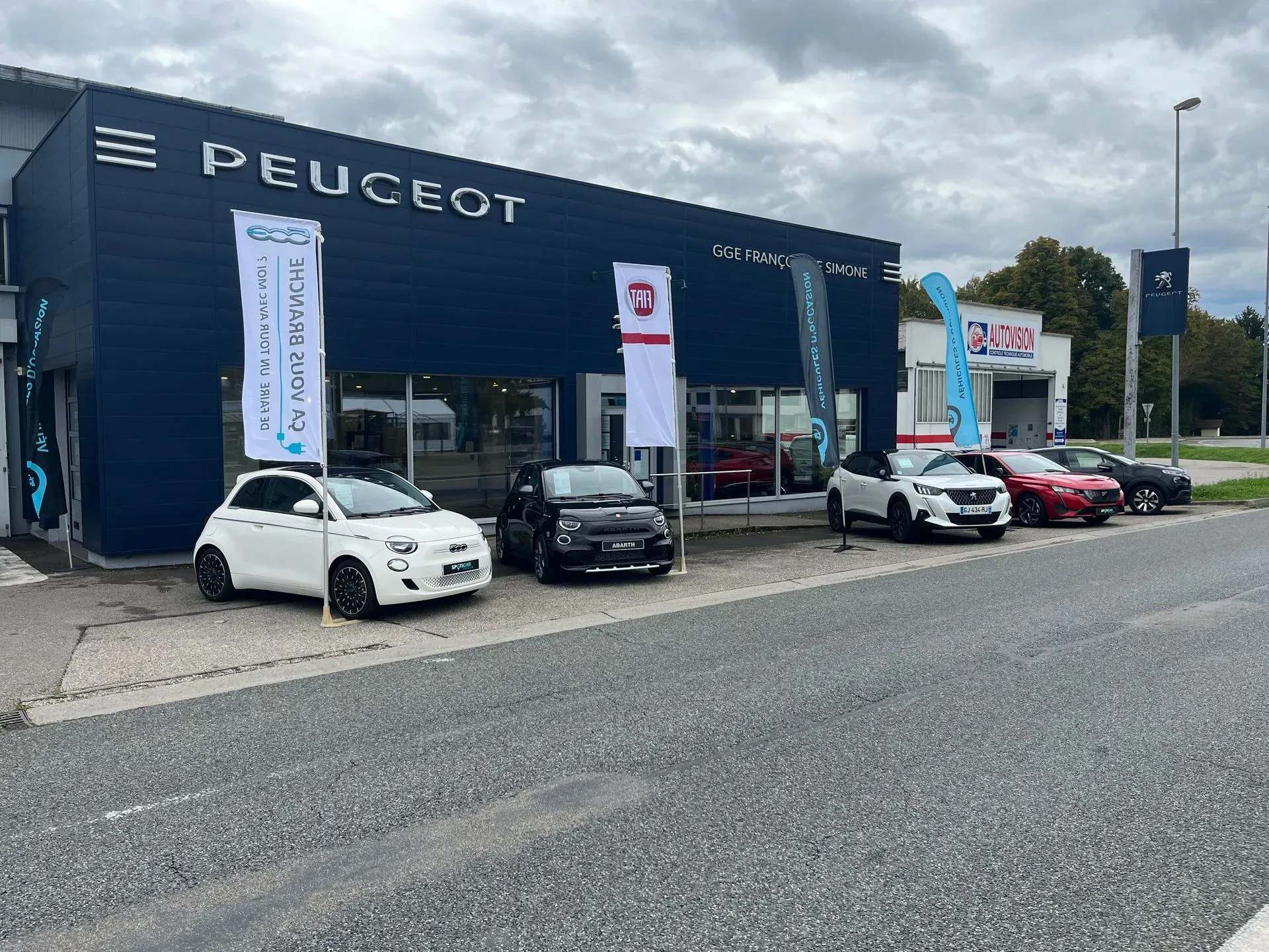 Façade bleue d'une concession Peugeot avec des drapeaux publicitaires et plusieurs voitures Fiat et Peugeot garées devant.