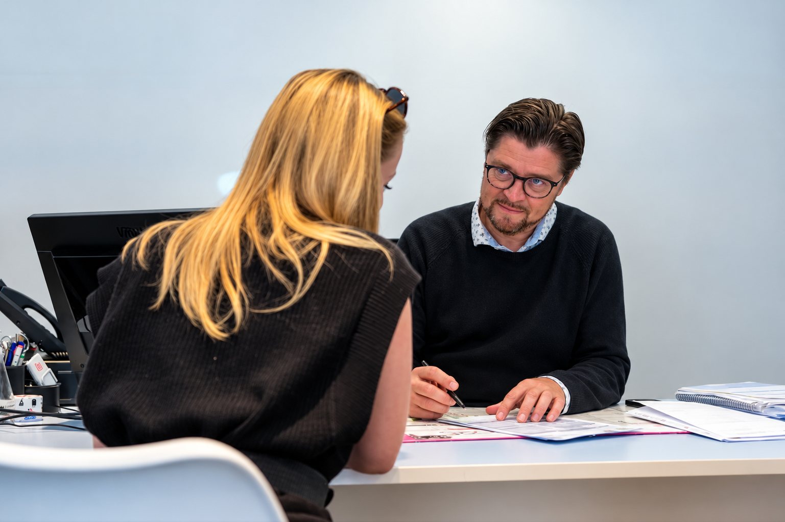 Un homme à lunettes en pull sombre discute avec une femme blonde vue de dos, assis à un bureau avec des documents devant eux.