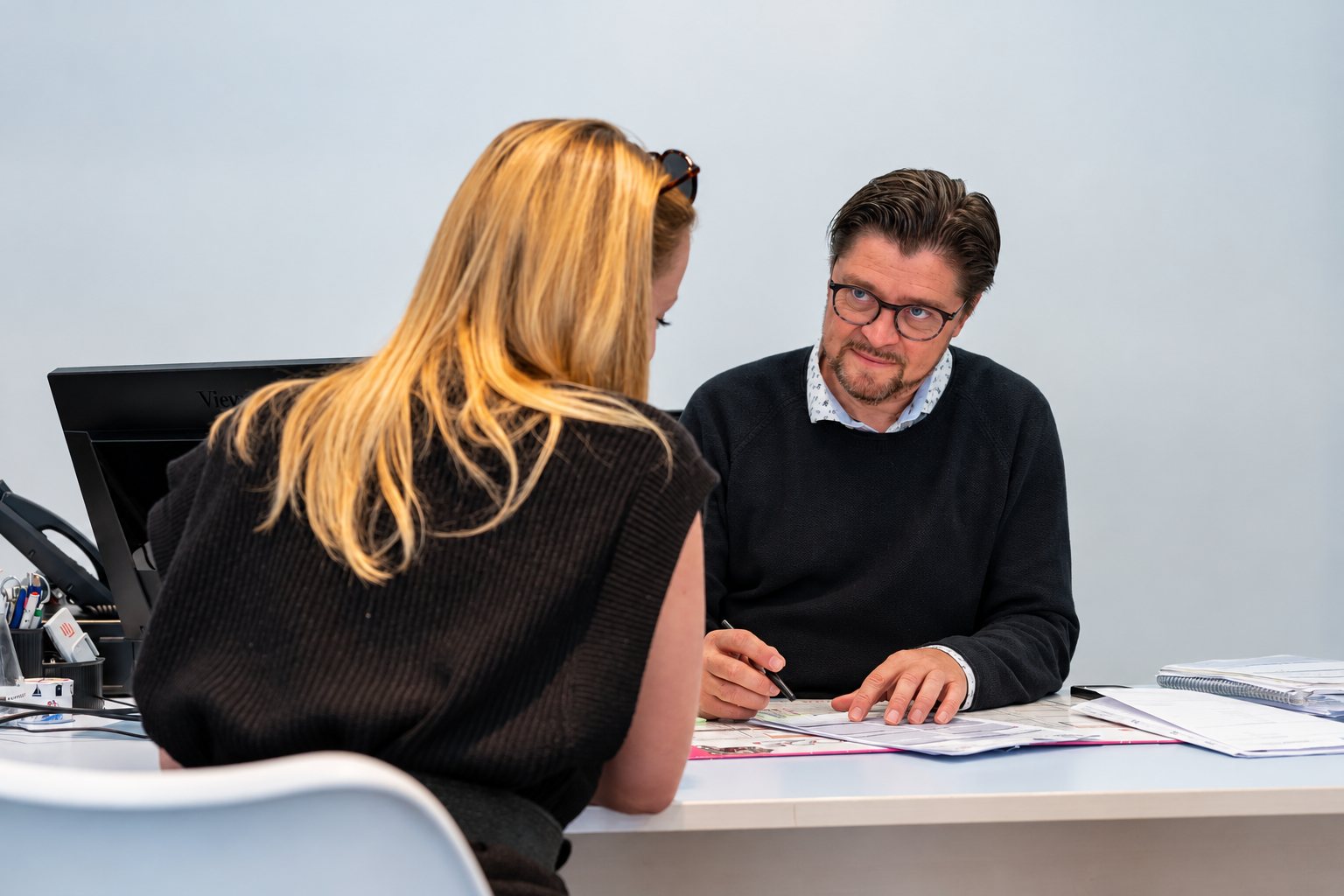 Un homme à lunettes vêtu d'un pull sombre discute avec une femme blonde vue de dos, assis à un bureau où sont posés des documents.