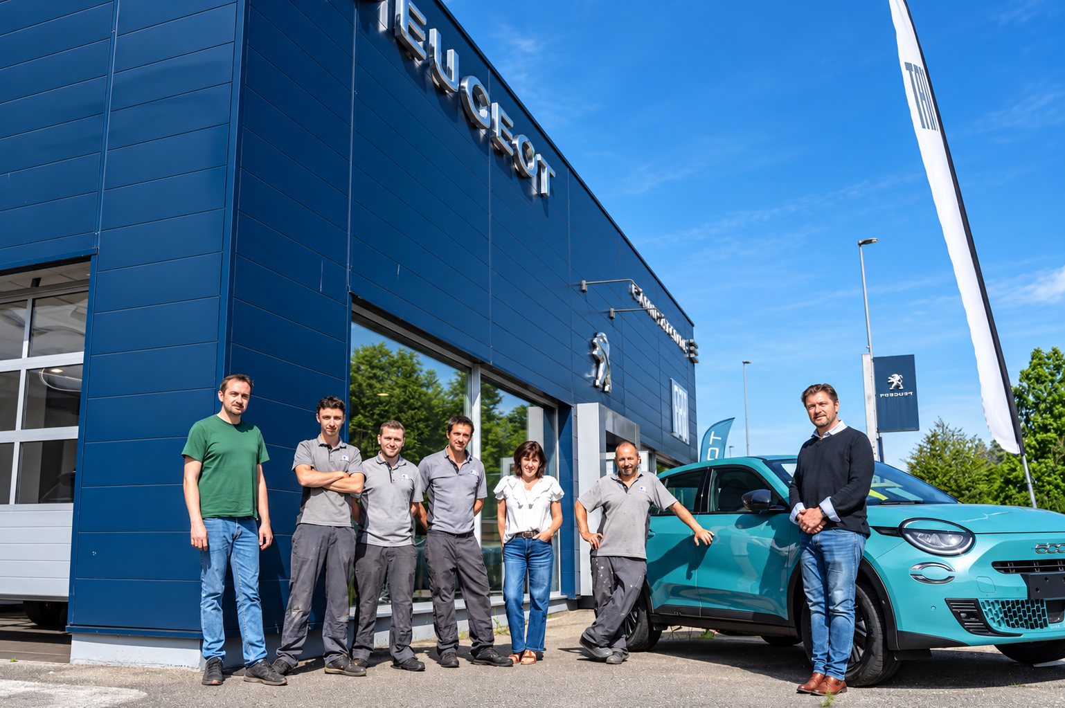 Sept membres de l'équipe d'un garage posent devant la façade bleue d'une concession Peugeot, à côté d'une voiture turquoise.
