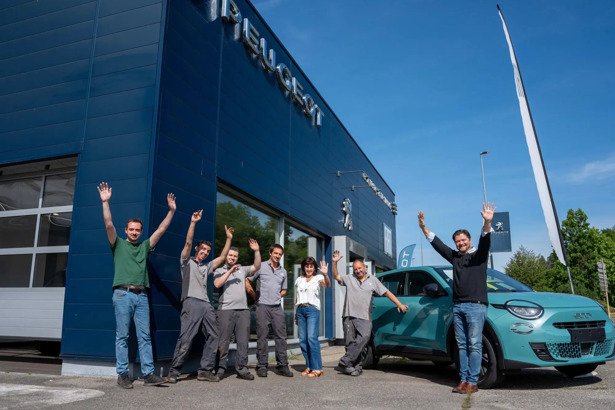 Sept membres de l'équipe d'une concession Peugeot lèvent les bras devant la façade bleue du bâtiment, à côté d'une petite voiture turquoise.