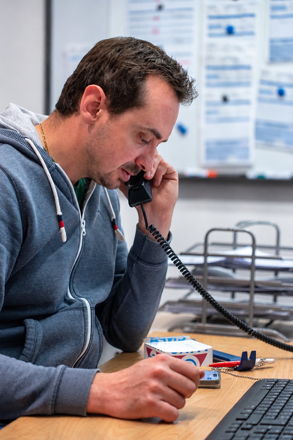 Un homme en sweat à capuche bleu gris parle au téléphone fixe à un bureau en bois, devant un tableau blanc couvert de notes.