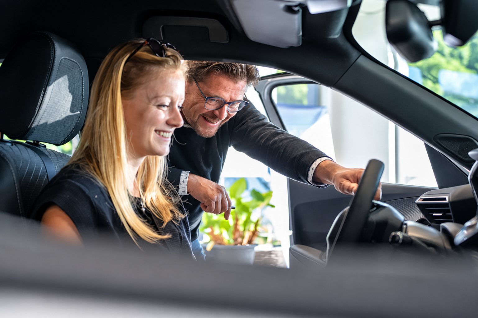 Un homme à lunettes se penche par la portière d'une voiture et montre le volant à une jeune femme blonde souriante assise au siège conducteur.